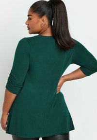 Vihreä pystyribattu tunika, jossa on kolmivartiset hihat ja levenevä helma. Pehmeä rakenne, jossa on hienovaraisia sivuärsytystietoja.