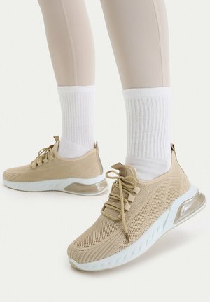 Beige gestrickte Sneakers mit weißen Sohlen und sichtbarem Luftkissen, getragen mit weißen, gerippten Socken und beigen Leggings vor einem schlichten Hintergrund.
