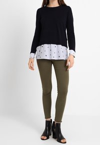 Mörkblå långärmad topp lagd över en randig skjorta med hjärt-mönster, i kombination med olivgröna snäva byxor och svarta sandaler med öppen tå.