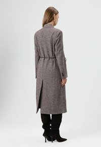 Lange grijze tweedjas met een tailleband, kraag en een achterzijdesplit, gedragen over zwarte kniehoge laarzen. Gladde textuur, op maat gesneden pasvorm.