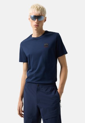 Młody mężczyzna z krótkimi blond włosami, noszący niebieskie okulary przeciwsłoneczne, granatową koszulkę i granatowe krótkie spodnie cargo, pozujący z jedną ręką w kieszeni.