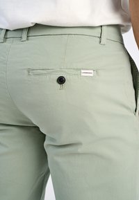 Pantalons vert clair en tissu doux, dotés d'une poche arrière avec un bouton noir et une étiquette blanche avec le nom de la marque.