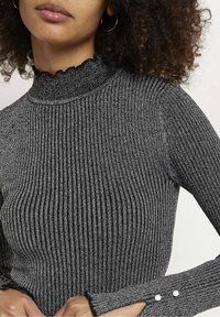 Grauer gerippter Rollkragenpullover mit einem gewellten Kragen, langen Ärmeln mit Knopfdetails und einem strukturierten, metallischen Fadendetail.
