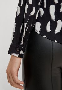 Blouse noire avec un motif de coups de pinceau blancs, dotée d'un poignet à boutons. Associée à un pantalon ajusté texturé noir.