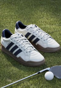 Λευκά παπούτσια γκολφ με μαύρες ρίγες πάνω στο γρασίδι δίπλα σε σκουπόξυλο και μπάλα γκολφ, υποδηλώνοντας γκολφ περίπτωση.