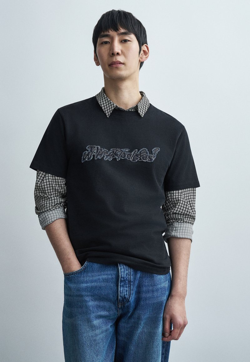 Μαύρο κοντομάνικο t-shirt με κεντημέν ο κείμενο, φορεμένο πάνω από γκρι και μαύρο καρό πουκάμισο. Μπλε τζιν τζιν με ανοιχτό πλύσιμο.