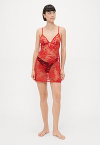 Woman standing barefoot wearing red sheer floral lace lingerie dress and black underwear, looking to the side against a plain background.