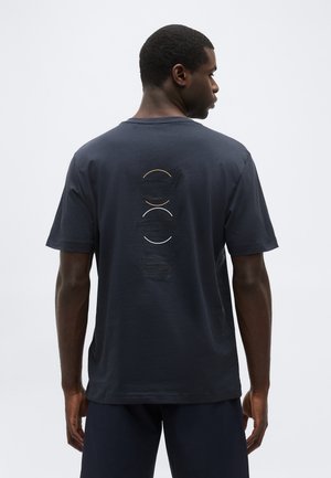 Férfi, aki sötétkék pólót visel, amelynek hátoldalán három dombornyomott kör alakú minta található narancssárga, fehér és fekete színekben, egyszínű háttér előtt áll.