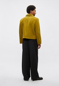 Volnena jopica gorčično rumene barve z ravnim ovratnikom, skrajšanim krojem in vidnimi šivi, v kombinaciji z širokimi črnimi hlačami in črnim obutvam.