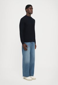 Mann står iført en svart teksturert genser, vide blå jeans og hvite plattform sneakers foran en helt hvit bakgrunn.