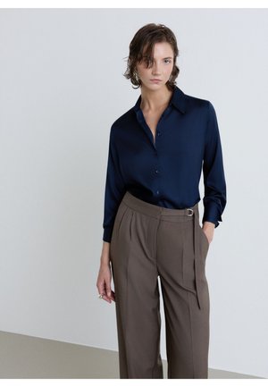 Femme aux cheveux courts et mouillés portant une chemise bleu foncé boutonnée rentrée dans un pantalon large marron taille haute avec une ceinture latérale.