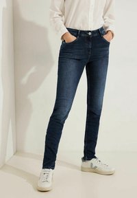Mörkblå, elastiska jeans med slimmad design, fem fickor och knapplås. Kombinerat med vita sneakers, modell stående.