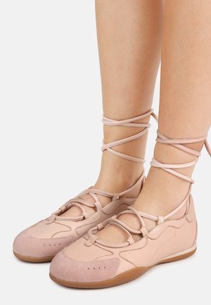 Pies con bailarinas de encaje color rosa pálido con cordones, tiras que se envuelven alrededor del tobillo y suelas marrón claro sobre un fondo blanco.