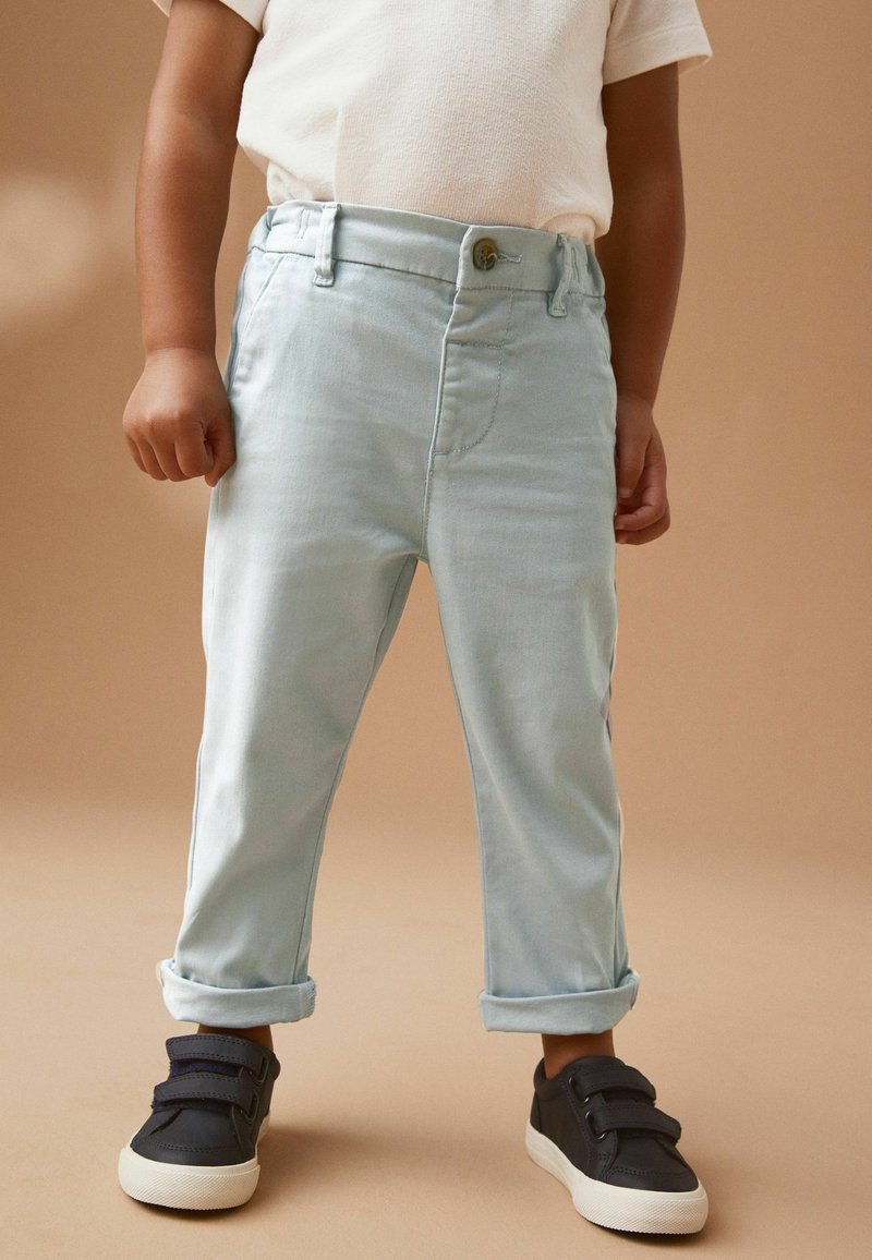 Bambino che indossa pantaloni azzurri risvoltati, camicia bianca e scarpe da ginnastica nere con chiusure a strappo, in piedi su uno sfondo neutro.