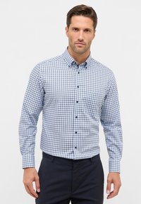 Gecarodeerde button-up blouse in wit, blauw en marineblauw. Heeft een kraag, lange mouwen en een enkele borstzak met blauwe knopen.