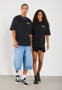 Divas melnas oversize T-krekli, kas uzvilkti divām modelēm, apvienoti ar ziliem džinsa šortiem un melnām potīšu zābaciņiem. T-krekliem ir grafisks teksta dizains.