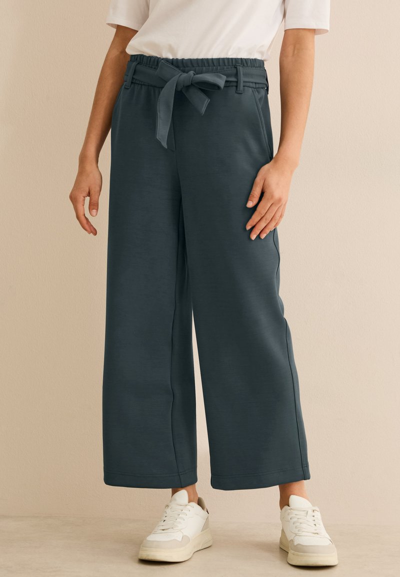 Pantalons larges en tissu vert foncé, dotés d'une taille élastique avec un lien, associés à des baskets blanches. Texture lisse et design décontracté.