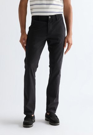 Mann trägt schwarze Slim-Fit-Hose, schwarze Schuhe und ein weißes kurzärmeliges Hemd mit horizontalen blauen Streifen, vor einem einfarbigen Hintergrund stehend.