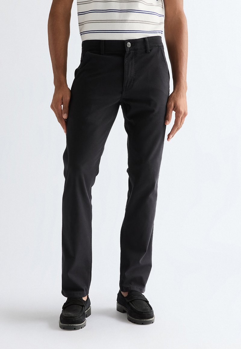 Mann trägt schwarze Slim-Fit-Hose, schwarze Schuhe und ein weißes kurzärmeliges Hemd mit horizontalen blauen Streifen, vor einem einfarbigen Hintergrund stehend.