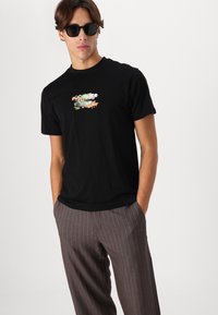 Tricou negru din bumbac cu un design grafic colorat pe față. Asortat cu pantaloni maronii cu dungi. Bărbat purtând ochelari de soare.