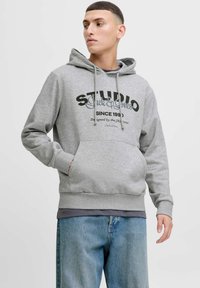 Šedá mikina s kapucí a přední kapsou, šňůrková kapuce a tištěný design textu. Nositá přes tmavé tričko a kombinovaná s světle modrými džíny.