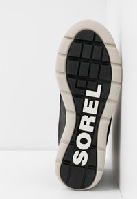 Schwarze Schuhsohle mit strukturiertem Muster, versehen mit einem weißen Sorel-Logo. Die Sohle ist dick und hat einen kontrastierenden beigen Rand.