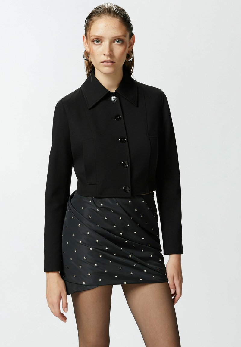 Crna skraćena jakna s ovratnikom i zatvaranjem na šest dugmadi, u kombinaciji s crnom suknjom koja ima metalne ukrase.