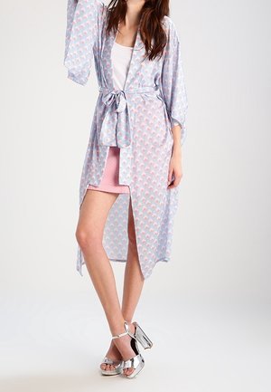 Femme portant une robe de chambre bleu clair à motifs nouée à la taille sur un haut blanc et une jupe rose, debout avec des sandales à talons hauts argentées.