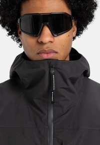 Black high-collar jacket with a zipper and textured fabric, paired with oversized black sunglasses. Smooth surface, minimal design features.