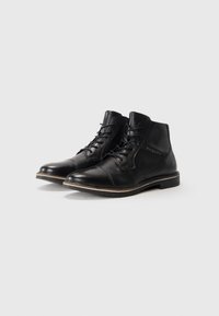 Botas de tobillo de cuero negro con cordones, con detalles de costura y un ribete marrón claro en la suela, con la marca "bugatti" en el lateral.