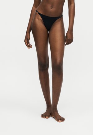 Partea inferioară a corpului unei persoane cu piele închisă la culoare, purtând slipi de bikini negri reglabili, stând desculță pe un fundal simplu deschis.