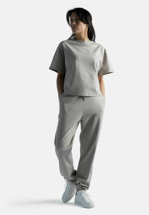 Nainen seisoo kädet taskuissa, yllään vaaleanharmaa väljä pusero, siihen sopivat jogger-housut ja valkoiset tennarit yksivärisellä taustalla.