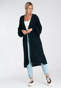 Długi, ciemnozielony kardigan dzianinowy o luźnym kroju, z otwartym przodem i dwiema przednimi kieszeniami. W połączeniu z jasnoniebieskimi dżinsami i casualowymi butami.