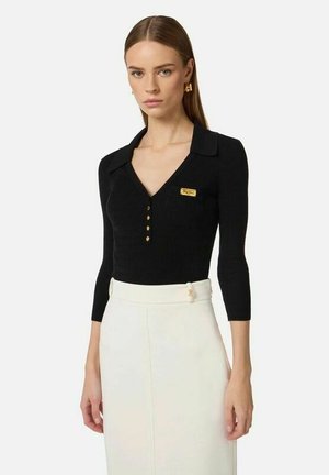 Donna che indossa un top nero a coste con bottoni dorati e una gonna bianca a vita alta su uno sfondo chiaro e uniforme.