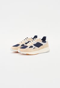Beige and navy sneakers featuring a suede and mesh upper, textured accents, and a contrasting white sole with an orange rubber outsole.