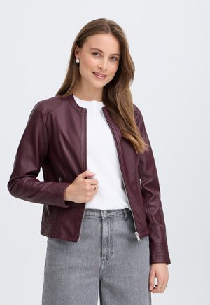 Femme aux longs cheveux bruns portant une veste en cuir bordeaux, une chemise blanche et un jean gris, posant et souriant devant un fond uni et clair.