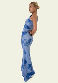 Femme portant une robe bleu clair à une épaule avec des motifs floraux bleu foncé, debout de profil, les mains dans le dos.