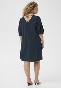 Vestido azul marino de manga abullonada con espalda en V y detalle de lazo. Tela suave y texturada, de longitud hasta la rodilla, con un corte holgado y costuras mínimas.