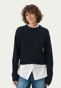 Navy blue cropped sweater with ribbed neckline and sleeves, featuring cable-knit detailing on the sides, layered over a white collared shirt.