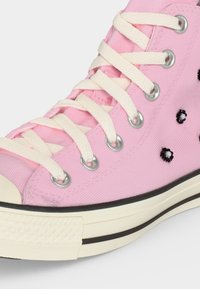 Κοντινό πλάνο ενός ροζ sneaker με ψηλό άνοιγμα, λευκά κορδόνια, μεταλλικά κρικάκια, ασπρόμαυρη κεντητή διακόσμηση με λουλούδια και λευκή λαστιχένια σόλα με μαύρη ρίγα.