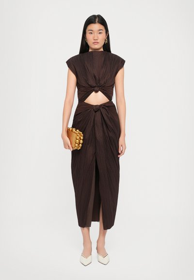 Rochie marron, pliată, cu mâneci scurte, detalii cu decupaj și design nodurat la talie. Modelul ține o poșetă rotundă cu accente aurii.