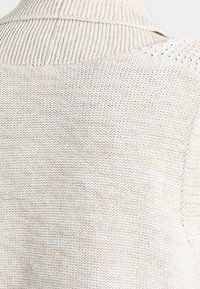 Ljust beige stickad tröja med ribbat krage, strukturerad mönster och öppet vävda detaljer på den övre delen av ryggen.