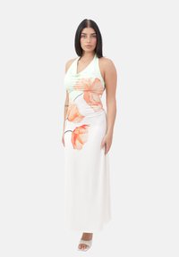 Femme aux cheveux bruns lisses portant une robe blanche sans manches avec de grandes imprimés floraux orange et des talons ouverts blancs, se tenant sur un fond blanc.