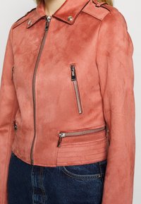 Jahodově růžová semišová bunda s límcem na patentky, předními kapsami na zip a žebrovanými detaily na bocích. Součástí jsou stříbrné kovové doplňky.