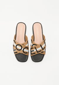 Sandales à glissière en tissu marron avec un design de brides croisées arborant des lettres noires et blanches. Semelle plate noire et marque discrète sur la semelle intérieure.