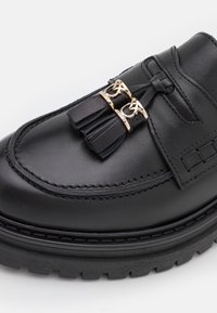 Mocassins en cuir noir avec des accents en métal doré, des glands et une semelle en caoutchouc texturée avec un motif de bande de roulement robuste.