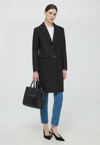 Cappotto di lana nero con una vestibilità su misura, due bottoni e tasche laterali, abbinato a jeans blu a vita alta e una borsa nera.