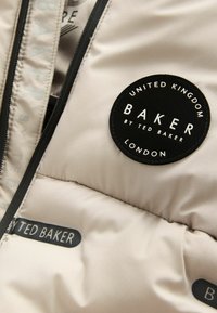 Puffermantel aus hellbeigem Stoff, ausgestattet mit einem runden schwarzen Logo mit "BAKER by TED BAKER", strukturierter Oberfläche und schwarzem Reißverschluss.