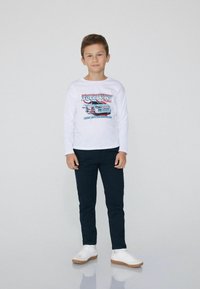 Chłopiec stoi, ubrany w białą koszulę z długim rękawem z grafiką samochodu, ciemne spodnie i białe sneakersy, na jednolitym jasnym tle.