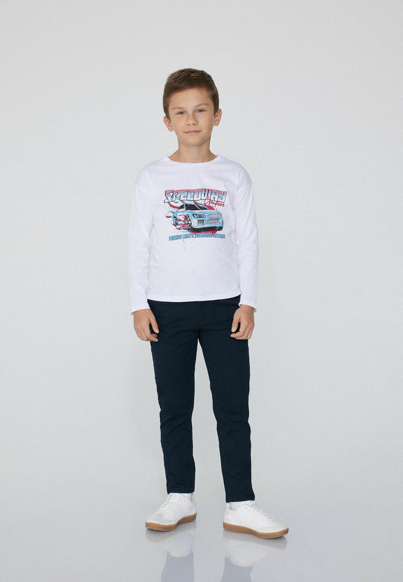 Chłopiec stoi, ubrany w białą koszulę z długim rękawem z grafiką samochodu, ciemne spodnie i białe sneakersy, na jednolitym jasnym tle.